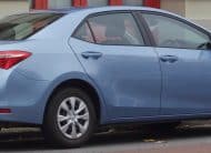 Blue Toyota Corolla sedan parked on a city street with red doors in the background and leaves on the curb.