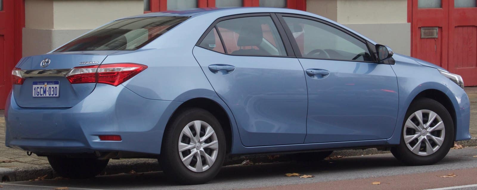 Blue Toyota Corolla sedan parked on a city street with red doors in the background and leaves on the curb.