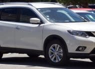 White Nissan SUV parked in a parking lot, front and side view showing the vehicle's grille and headlights.