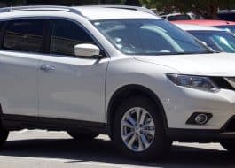 White Nissan SUV parked in a parking lot, front and side view showing the vehicle's grille and headlights.