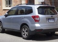 Silver Subaru SUV parked in a lot, viewed from the rear three-quarter angle beside a brick building with a gated area behind it.