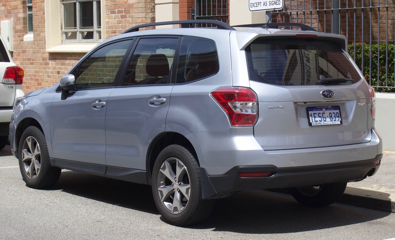 Silver Subaru SUV parked in a lot, viewed from the rear three-quarter angle beside a brick building with a gated area behind it.