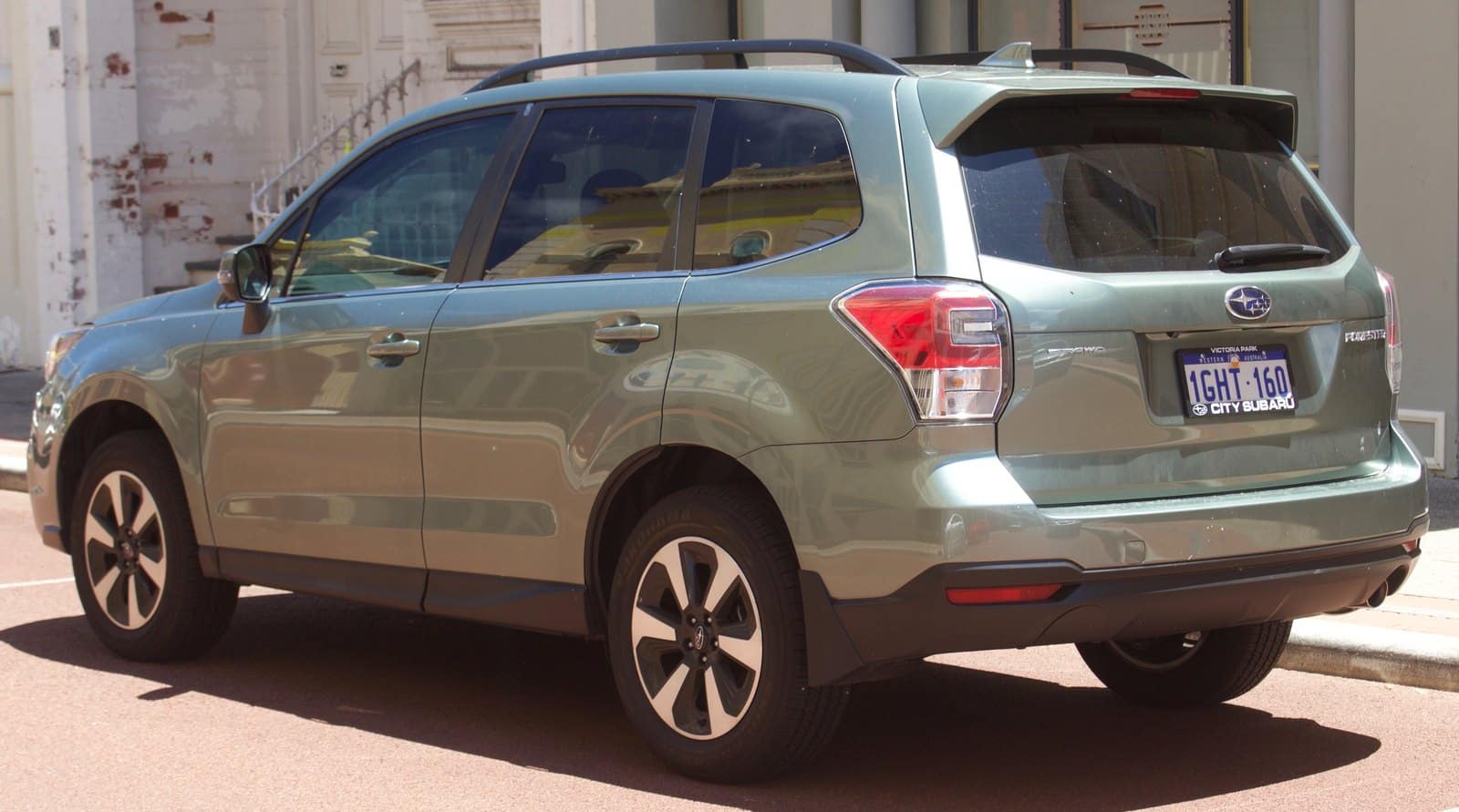 Rear three-quarter view of a light blue Subaru Forester SUV parked on a city street, with roof rails and visible license plate balance on the rear.