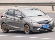 Gray Honda hatchback driving on a multi-lane highway with a blue license plate and a guardrail in the background, motion blur suggesting speed.