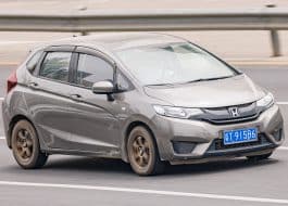 Gray Honda hatchback driving on a multi-lane highway with a blue license plate and a guardrail in the background, motion blur suggesting speed.