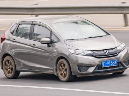 Gray Honda hatchback driving on a multi-lane highway with a blue license plate and a guardrail in the background, motion blur suggesting speed.