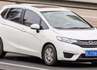 White Honda hatchback driving on highway, front grille and blue license plate visible