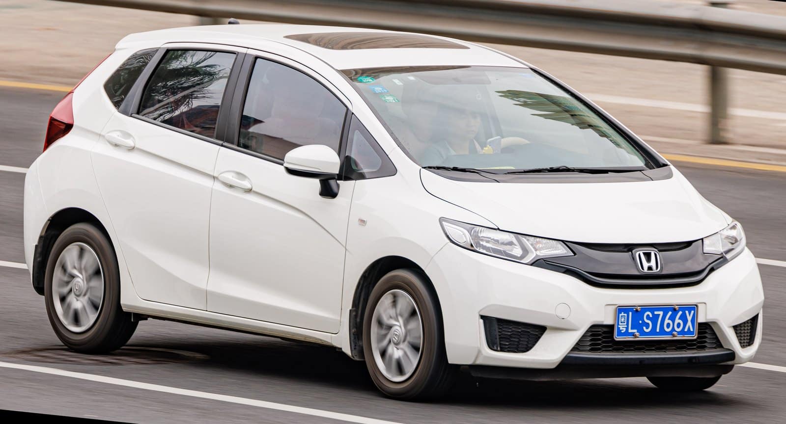 White Honda hatchback driving on highway, front grille and blue license plate visible