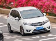 White Honda minivan driving on a city street with pink flowering bushes in the background. Front license plate visible (blue).