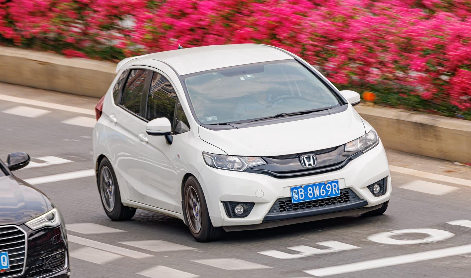 White Honda minivan driving on a city street with pink flowering bushes in the background. Front license plate visible (blue).