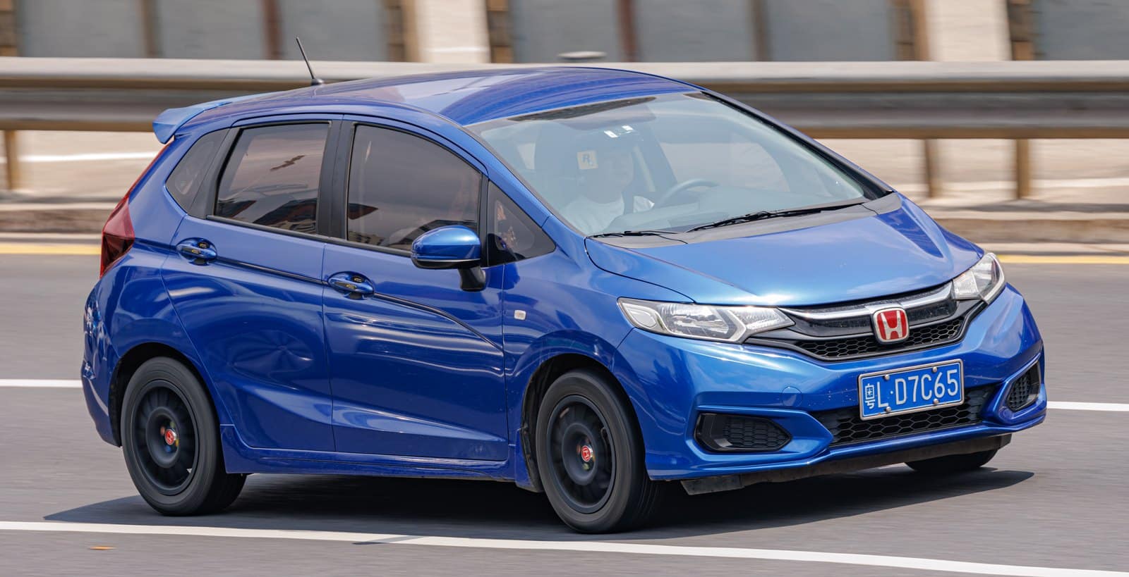 Blue Honda compact car driving quickly on a highway, license plate L D7C65, with a blurred guardrail background.