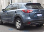 Blue Mazda CX-5 SUV parked on a city street, shown from the rear three-quarters with license plate visible.
